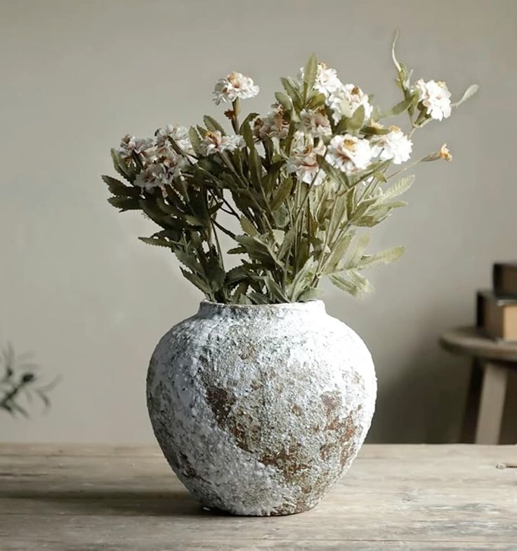 "Rustic ceramic vase with a weathered, textured finish, holding a bouquet of delicate white flowers and greenery, placed on a wooden table with a soft, neutral background."