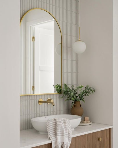 “Modern bathroom vanity with a white countertop, vessel sink, brass fixtures, and a gold-framed arched mirror. A vase with greenery adds a touch of nature, complemented by soft lighting.”