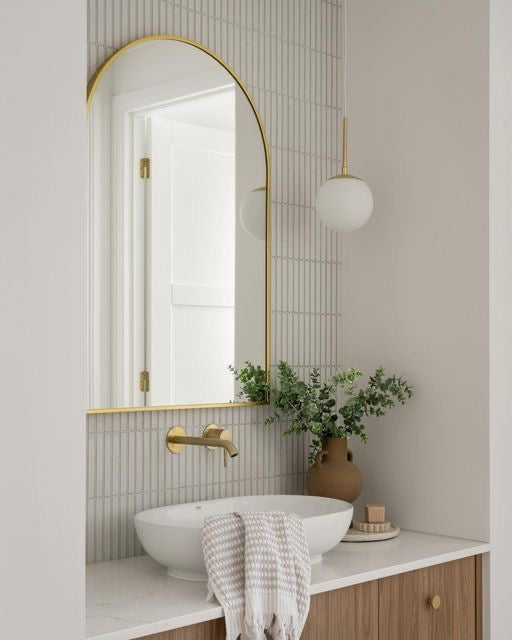 “Modern bathroom vanity with a white countertop, vessel sink, brass fixtures, and a gold-framed arched mirror. A vase with greenery adds a touch of nature, complemented by soft lighting.”