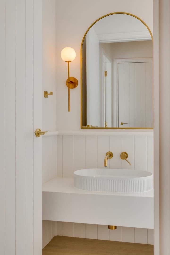 “Minimalist bathroom with a white shiplap wall, ribbed ceramic vessel sink, and brass faucet fixtures. An arched gold mirror and a brass sconce light provide an elegant finish.”
