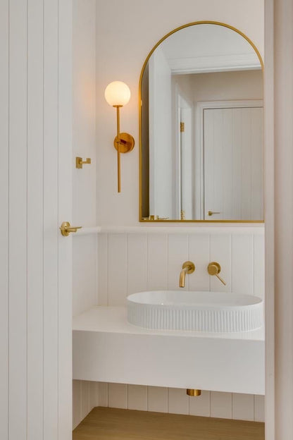 “Minimalist bathroom with a white shiplap wall, ribbed ceramic vessel sink, and brass faucet fixtures. An arched gold mirror and a brass sconce light provide an elegant finish.”