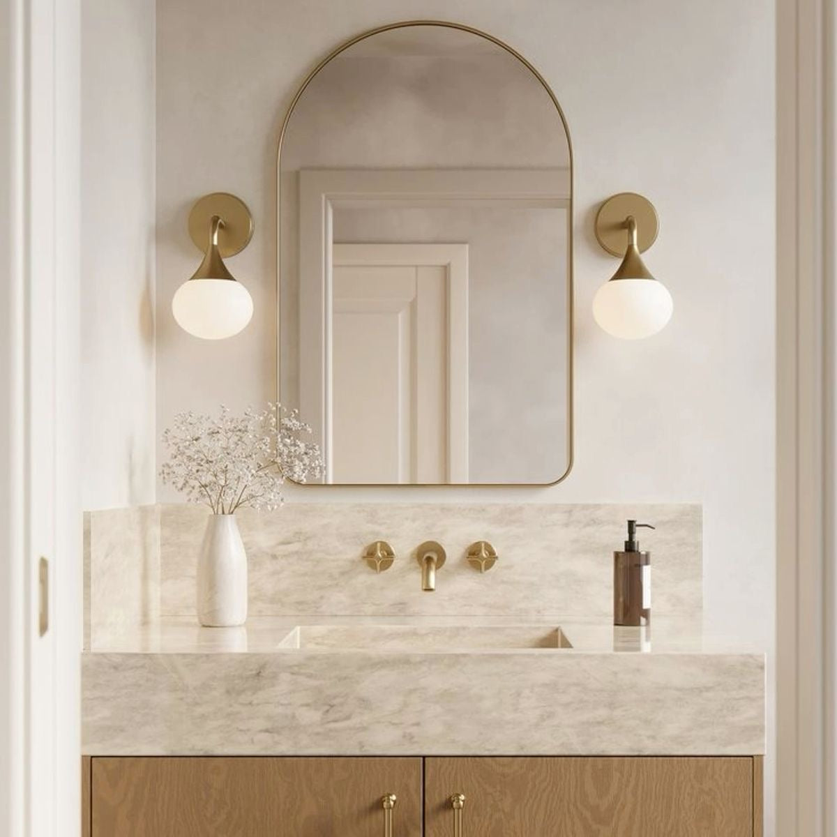 “Sophisticated bathroom design featuring a marble vanity, brass fixtures, and an arched mirror framed in gold. Wall sconces with round glass globes and a vase with delicate flowers add a luxurious touch.”