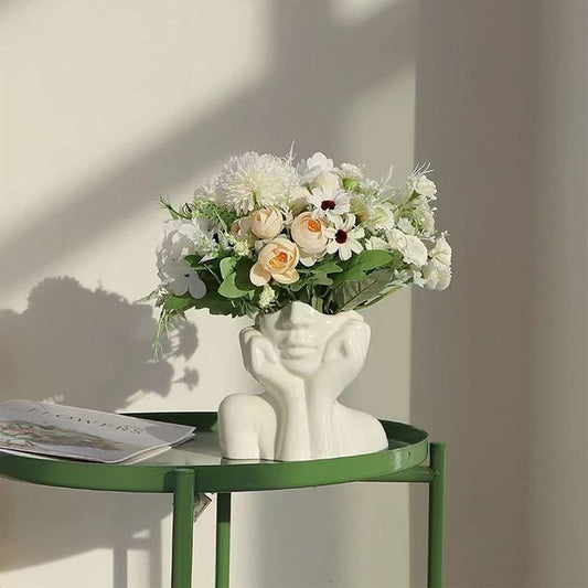 A charming white ceramic female face vase with a rustic green round table, showcasing an arrangement of white and blush floral blooms in a serene and sunlit corner.