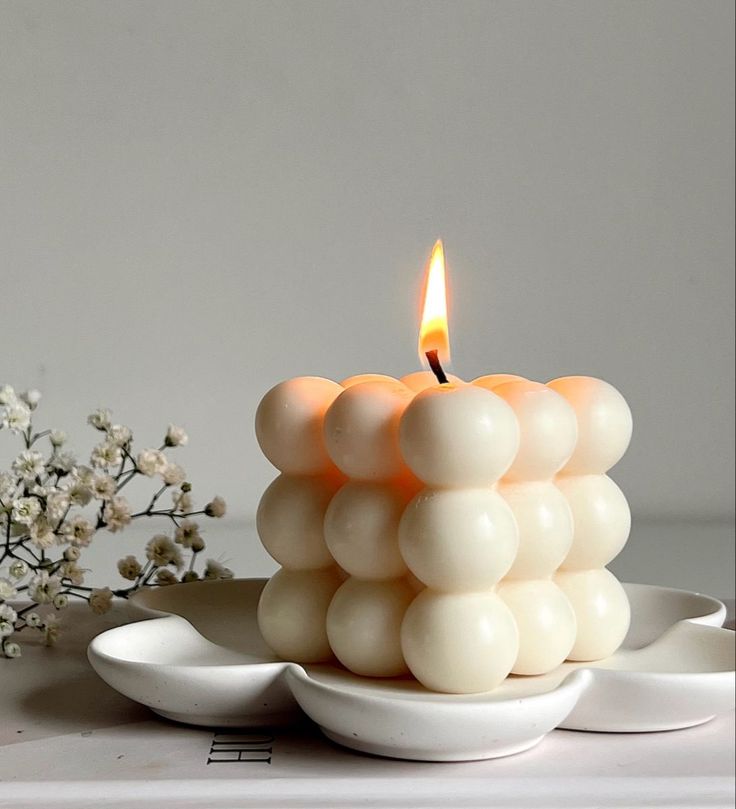 "A glowing bubble candle with a single flame, displayed on a white ceramic dish with delicate baby's breath flowers in the background."