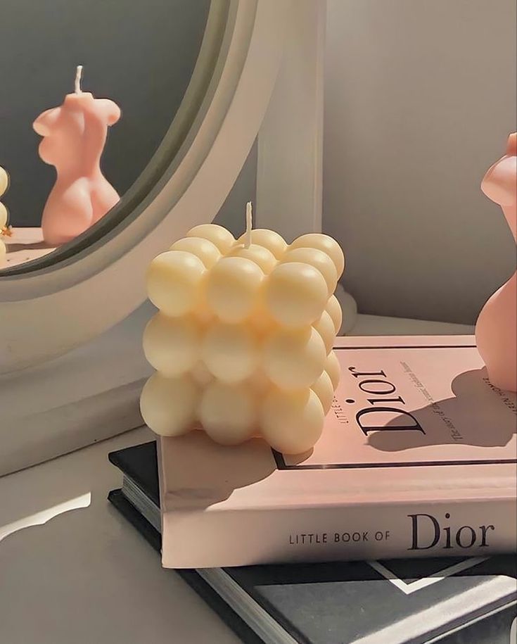 "Cream bubble candle styled atop a stack of chic books, including 'Little Book of Dior,' with soft lighting and a pink feminine torso candle reflected in a mirror nearby."