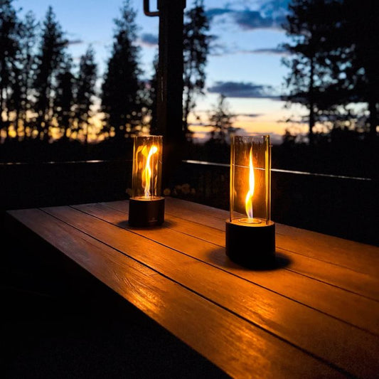 "Two elegant glass flame lanterns on a wooden table, softly glowing against the backdrop of a serene evening sky with silhouetted trees and a colorful sunset."