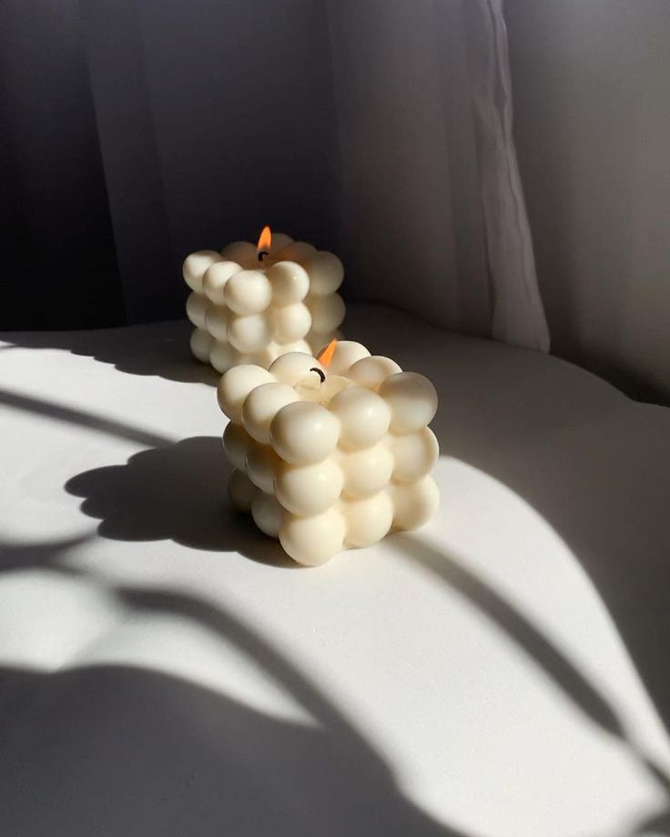 "Two lit bubble candles in creamy white tones, casting soft shadows in natural light, placed on a smooth white surface near sheer curtains."
