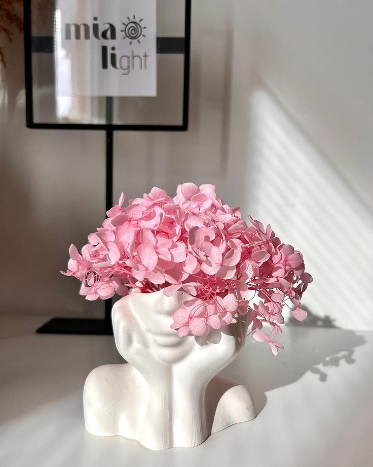 A minimalist pink-themed decor featuring a white ceramic female face vase holding delicate pink hydrangeas, styled on a modern desk with natural sunlight casting soft shadows.