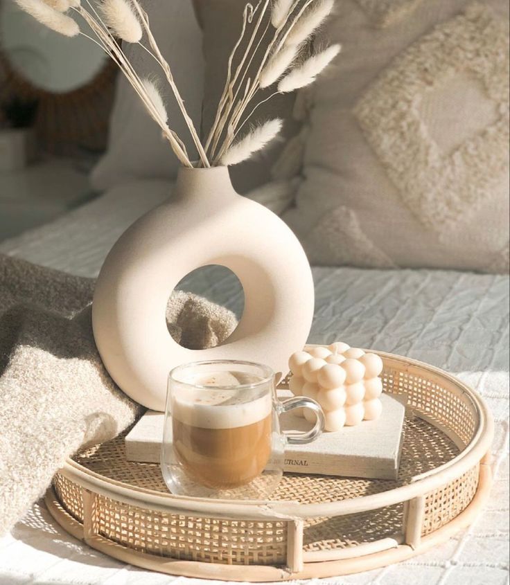 "Cozy tabletop setup featuring a circular ceramic vase with pampas grass, a bubble candle, and a latte served on a rattan tray, placed on a soft, beige-toned bed."