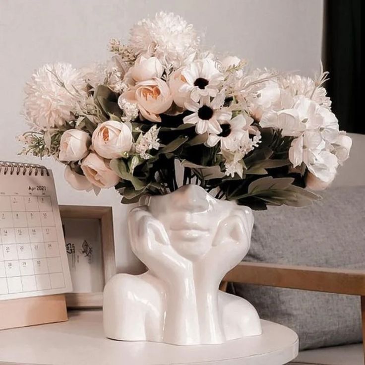 A chic white ceramic vase shaped like a thoughtful female face, filled with elegant white and blush pink flowers, sitting on a white table next to a calendar and minimal decor.