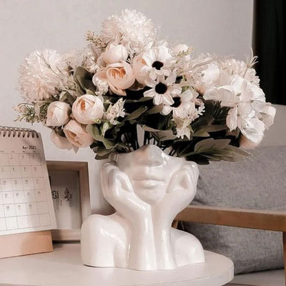 A chic white ceramic vase shaped like a thoughtful female face, filled with elegant white and blush pink flowers, sitting on a white table next to a calendar and minimal decor.